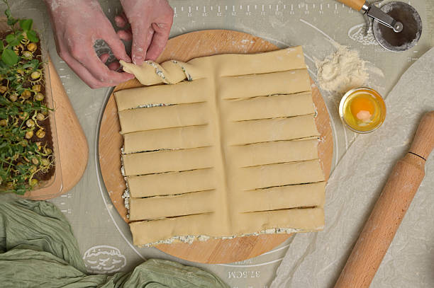 istock Top view woman's hands twists dough strips on wooden board. Cutter, egg, green towel, microgreens sprouts on kitchen table. Home cooking baking puffs 2207842833