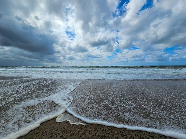 Waves crash onto a sandy beach beneath a dramatic cloudy sky with patches of blue. The photo captures the raw power and beauty of nature by the sea, evoking a sense of calm and energy.