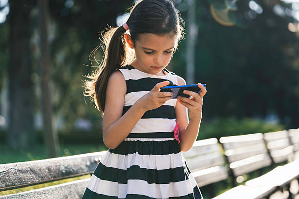 A girl stands alone in a sunny park, focused intently on her smartphone, embodying modern childhood activities and the engagement of young minds in technology.