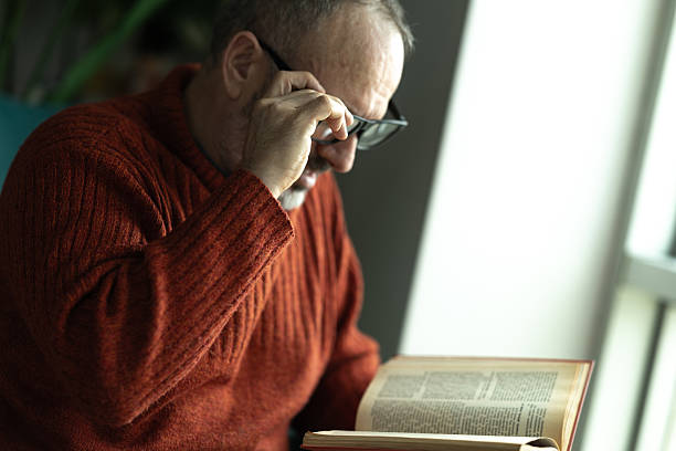 Mature man peering through his glasses while reading a book near a window