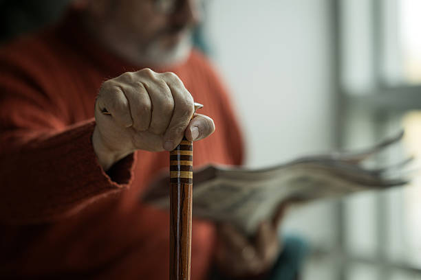 Close-up of elderly man's hand gripping walking cane while reading newspaper, emphasizing themes of aging, mobility, and daily life