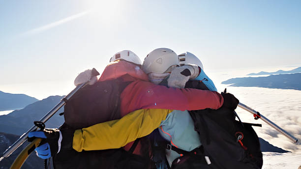Huddle of mountaineers on peak, embracing in camaraderie, overlooking snowy mountains and clear sky, conveying team spirit and relaxation.