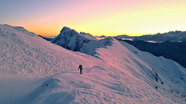 Snowy mountain range at sunset, with a lone hiker trekking uphill, evokes a sense of adventure and tranquility.