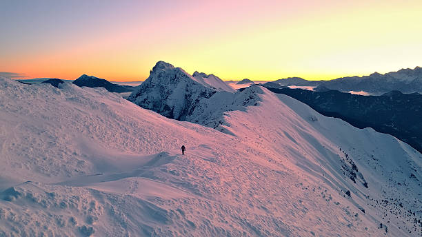 Breathtaking panoramic view of a snow-covered mountain range at sunset, with a lone climber ascending the slope, evoking a sense of adventure and tranquility.