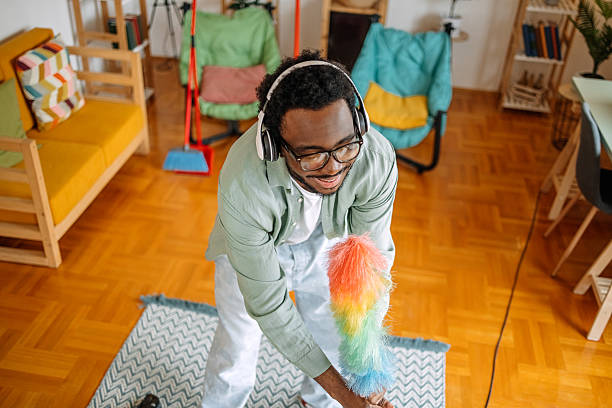 Mid adult man cleaning dust from a houseplant with a feather duster, savoring his weekend activity in a modern, cozy living room filled with natural light and comfortable furniture