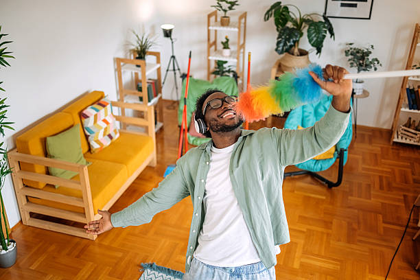 Young black adult listening music in living room while dusting and acting like he is a singer using dust cleaner like it is a microphone