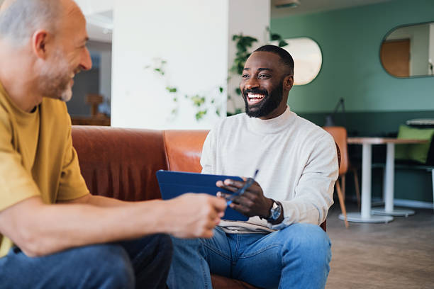 Two Smiling Businessmen Engaged in Conversation While Sitting in a Modern Office Lounge Two men enjoy a friendly chat in a cozy office lounge, fostering a professional yet relaxed atmosphere. Their interaction exudes positivity and collaboration, reinforcing themes of teamwork and communication. office buddies stock pictures, royalty-free photos & images