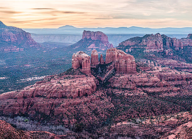 Aerial view of Cathedral Rock in Sedona, Arizona, captured at dawn. The warm glow of sunrise bathes the red rock formations in soft pastel hues while distant mesas and buttes fade into blue layers across the horizon. This sweeping composition highlights the serene beauty and vastness of the American Southwest.