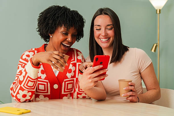 Two joyful women happily sharing a delightful moment while using a smartphone to watch a funny video and enjoying delicious drinks in a cozy indoor setting with warm smiles and laughter