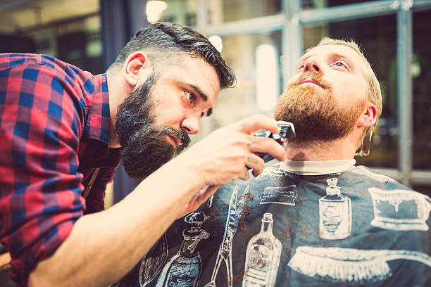 Barber carefully trimming a bearded customer's beard in a modern barbershop during nighttime, creating a clean and stylish look.