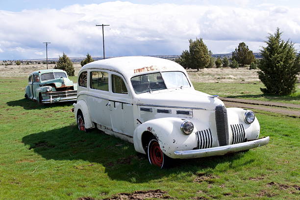 Old white sedan car abandoned in a tall grassy field with trees and scattered vehicles in Saskatchewan, Canada.
