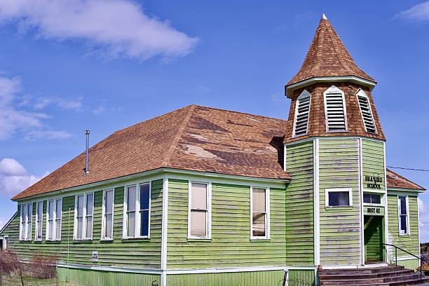 A weathered yet charming historic schoolhouse in Shaniko, Oregon, painted in faded green with a distinctive bell tower. The old wooden structure shows signs of aging, with peeling paint and a worn roof, yet it stands as a testament to the town's past.