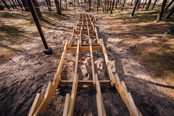 Wooden staircase under construction on a forested sandy slope, supported by metal posts, with caution tape and trees surrounding the site.