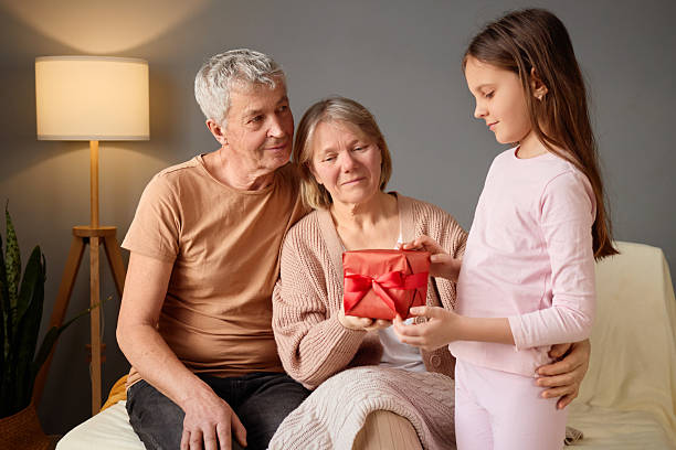 istock Grandparents sit together on a couch while their granddaughter presents a bright red gift with a bow. Warm lighting creates a cozy atmosphere in the living room 2207901964