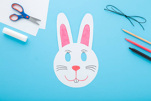 White smiling rabbit paper mask on light blue table background. Pastel color. Closeup. Creating easter decoration for kid. Top down view.