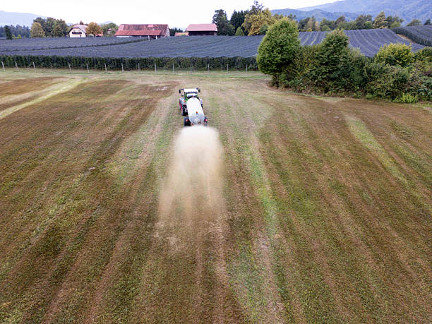 Aerial view of a tractor spraying fertilizer on a field. The landscape features rows of crops, trees, and distant barns under a clear sky.