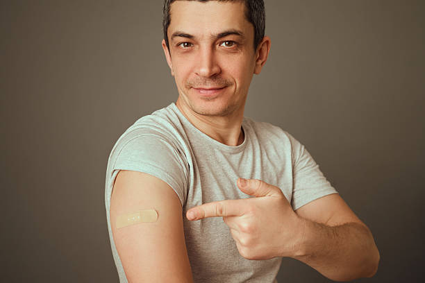 istock A man in a gray shirt smiles confidently while pointing to a bandage on his arm after getting vaccinated. The setting features a neutral background that emphasizes his expression 2207905929