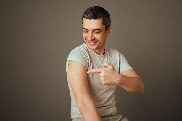 istock A man in a gray t-shirt proudly points to his arm where a small bandage is visible, indicating he has just received a vaccination. His expression is positive and satisfied 2207905933