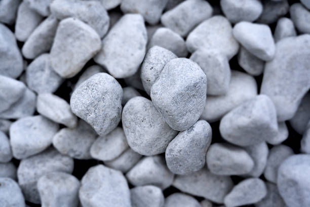 The close-up image shows a pile of small rocks and pebbles. Stones of various sizes and colors, including blue, black, and white. In the center of the image is a large round stone with a rough surface. The stone is covered with small white specks that give it texture. To the left of the stone are two small orange objects that look like pebbles, one on top of the other. The background is blurred, making the stones the center of attention.