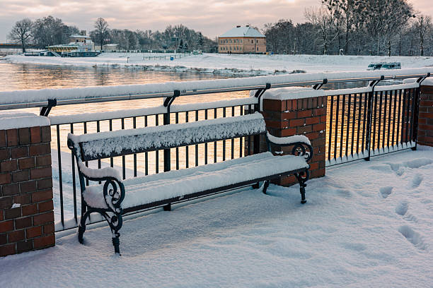 A bench covered with beautiful fluffy snow against the backdrop of a river. Dawn on a winter river.