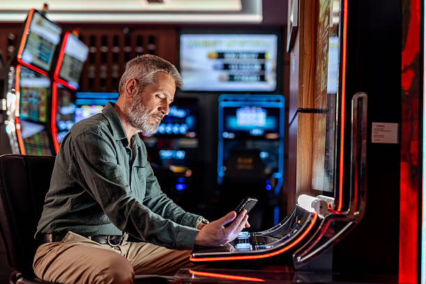 A mature man interacts with his smartphone while seated at a modern casino slot machine, bathed in ambient neon lights, evoking a scene of focus and leisure within a gaming environment.