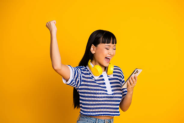 istock Joyful Woman Celebrating Achievement While Using Smartphone Against a Bright Yellow Background 2207913143