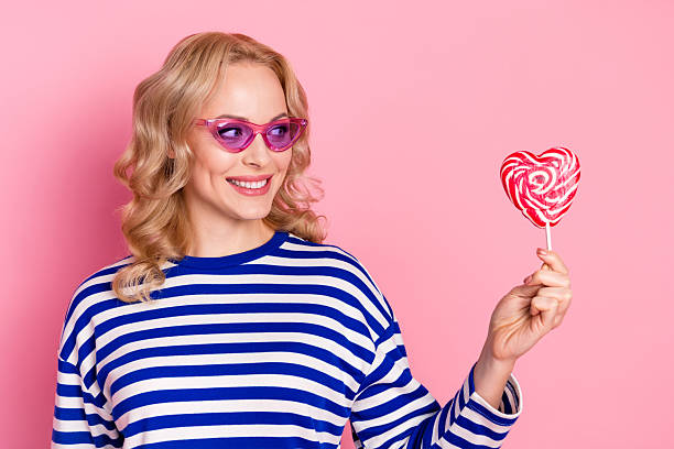 istock Charming blonde woman in a stylish striped pullover holding a heart shaped candy, smiling cheerfully against a cheerful pink background 2207913915