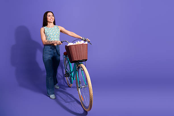 istock Young woman in casual stylish attire smiling while pushing a modern bicycle with a wicker basket against a purple backdrop 2207914830
