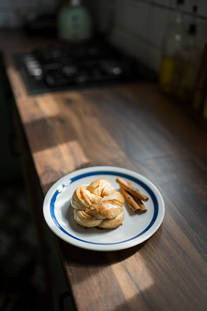 Freshly baked cinnamon roll with cinnamon sticks sits on a plate in a sunlit modern kitchen, ready to be enjoyed