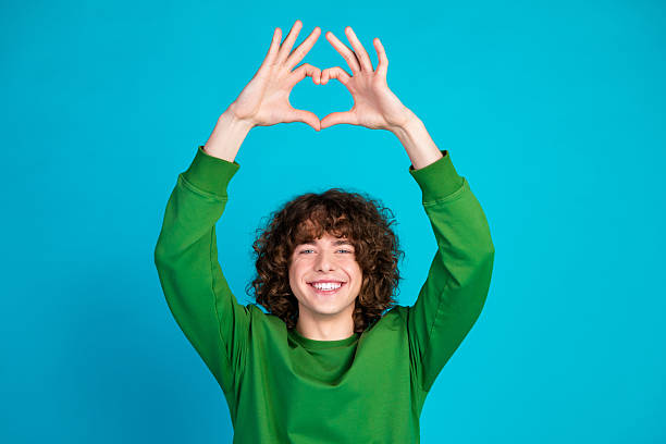 istock Smiling Young Man in Green Sweatshirt Making Heart Gesture with Hands Against Blue Background 2207915044