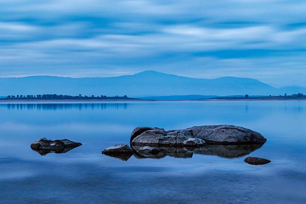 Serene lake with rocks in the foreground and distant mountains under a cloudy blue sky at dusk.