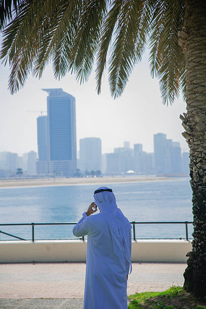 Portrait of middle eastern businessman with traditional emirati clothes, smiling, speaking with smartphone and looking away from the camera against Dubai cityscape, UAE.