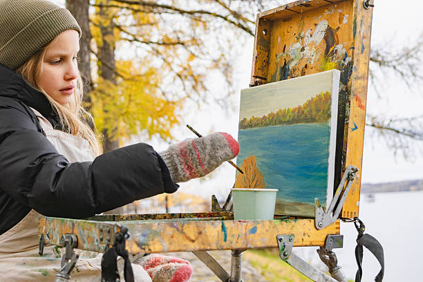A young girl dressed in warm clothing and mittens paints a serene river landscape on a small canvas using a portable easel in an autumn outdoor setting, capturing the beauty of nature through art