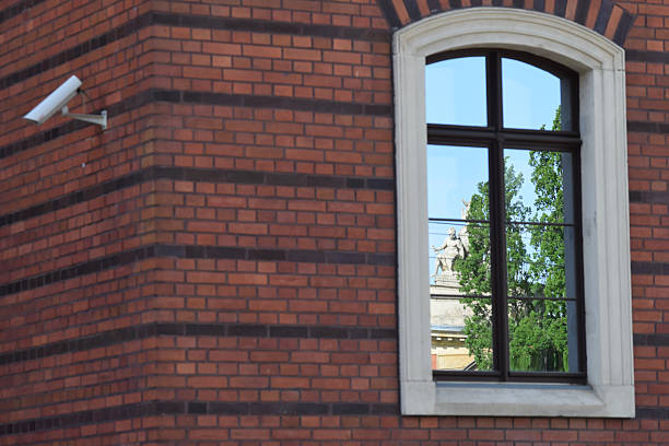 The facade of a red brick building features a window divided into square panes, reflecting a powerful and clear image of a monument from the opposite building. The monument depicts a man appearing as if he is escaping through the window. To the left, a security camera is mounted on the wall, adding a modern contrast to the historic architecture.