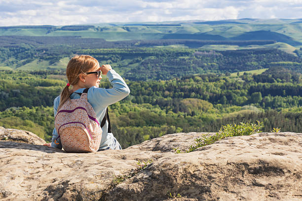 A young girl wearing a floral backpack sits on a rocky cliff and shades her eyes with her hand while looking out over a vast green valley and forested mountains during a sunny spring day