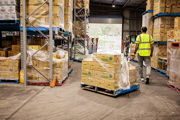 Professional warehouse worker in high-visibility gear uses a pallet jack to move merchandise between metal racks in a spacious storage room.