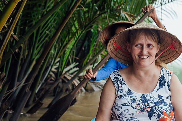 A European tourist sits smiling on a canoe , as she is rowed along by an anonymous guide sat behind her. This lone traveller is boating beside the river edge palm trees. The two boat occupants sit facing forward and both wear traditional triangular palm frond hats.