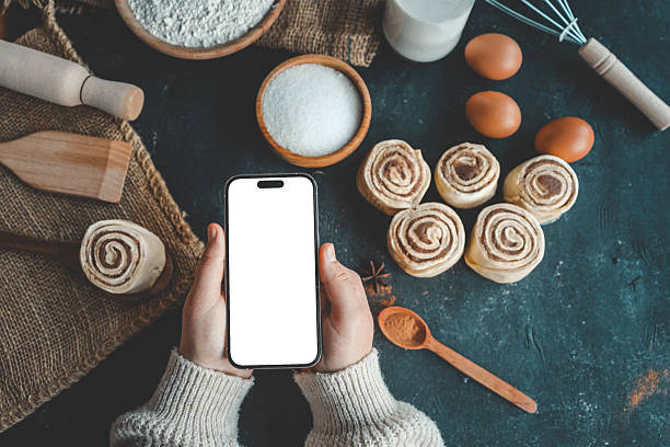 istock Hands holding smartphone with blank screen near raw cinnamon rolls, flour, sugar, eggs, and kitchen tools. Baking ingredients on dark table, top view 2207948176