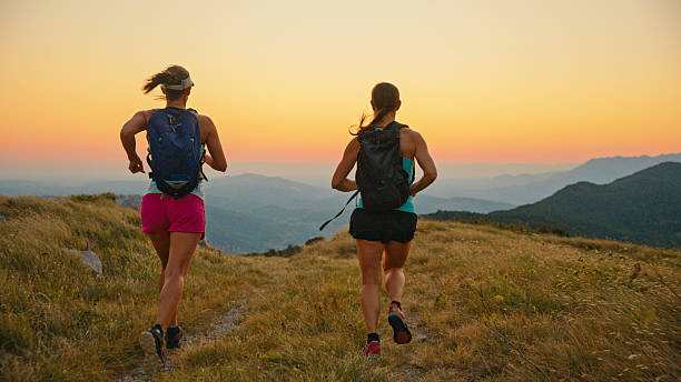 Two Women Run Together on a Grassy Mountain Slope,Moving Away From the Camera Towards a Breathtaking Panoramic View of Distant Peaks Under a Colorful Sunset Sky. The Image Captures the Effort and Beauty of Trail Running in a Stunning Natural Landscape During the Golden Hour