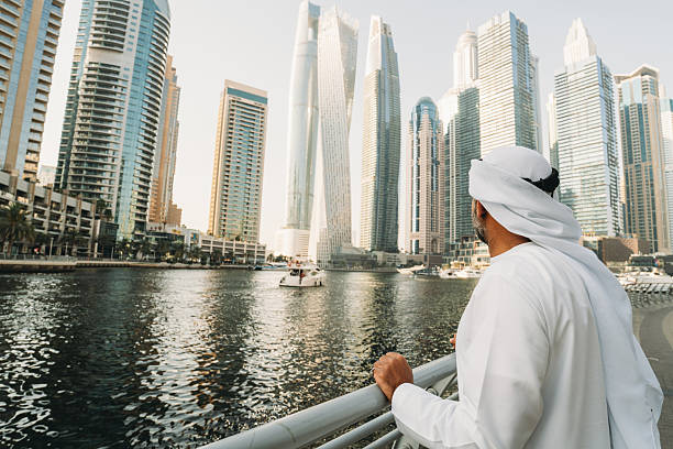 Emirati businessman in traditional clothing is looking at the Dubai Marina skyline from a bridge