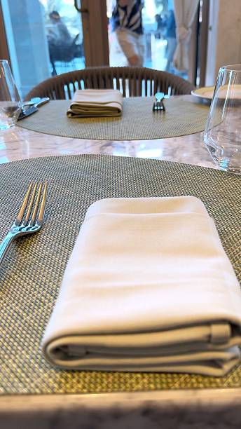 istock Round table holds folded napkins and flatware atop woven mat, clean setup stands ready for service while daylight filters across glass and chairs in soft elegant restaurant tone. 2207980492