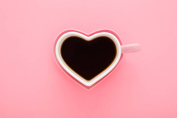 Heart shape white cup and pink saucer. Fresh black coffee cup on light pink table background. Pastel color. Closeup. Top down view.