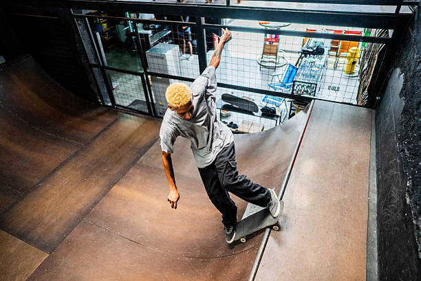 A skateboarder with blond hair shifts their weight during a tight maneuver in an indoor skatepark, framed by rails and warm lighting.