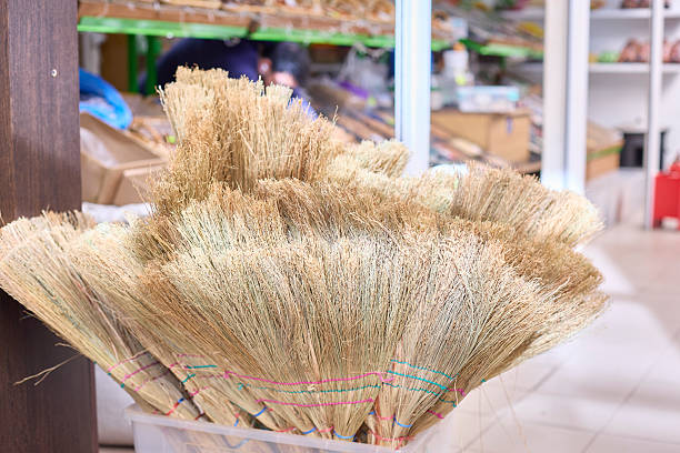 Close-up of natural straw brooms neatly arranged in a basket inside a store. The scene highlights their texture and craftsmanship, with shelves of goods in the background.