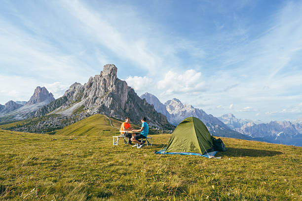 Two men sit at a table next to a tent in Passo Giau, surrounded by the dramatic scenery of the Dolomites under a blue sky.