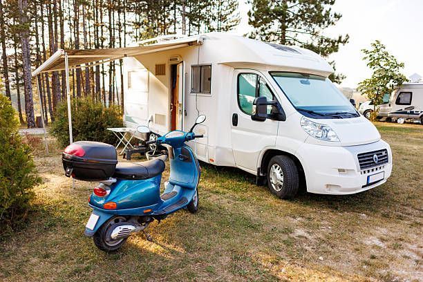 White Camper van and blue motor scooter parked at a campsite on sunny day in a wooded outdoor setting