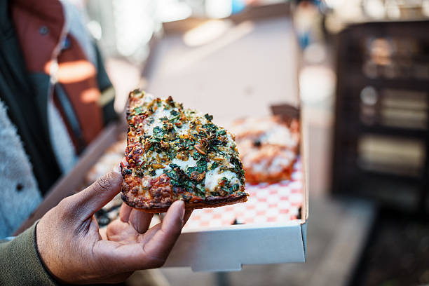 Cropped view with selective focus on a gourmet slice of pizza in the hand of an unrecognizable Filipino man as he eats takeout street food while exploring San Francisco, California.
