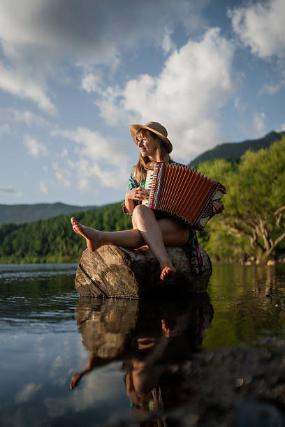 Young woman musician playing accordion while sitting on a log in a lake with mountains in the background