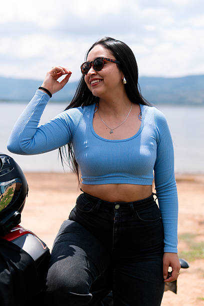 Fashionable young woman adjusts her sunglasses while leaning on her motorcycle, enjoying the scenic view of a lake