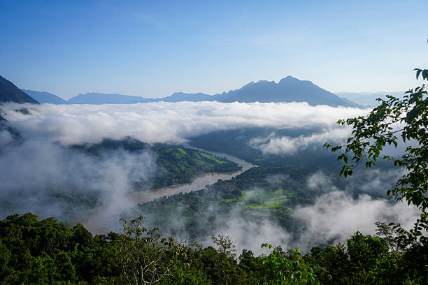 A tropical river in Nong Khiaw, Laos, at dawn, reflecting the soft hues of the early morning sky. Layers of clouds drift above the calm water, while mist lingers over the distant jungle-covered hills, creating a tranquil and atmospheric landscape.